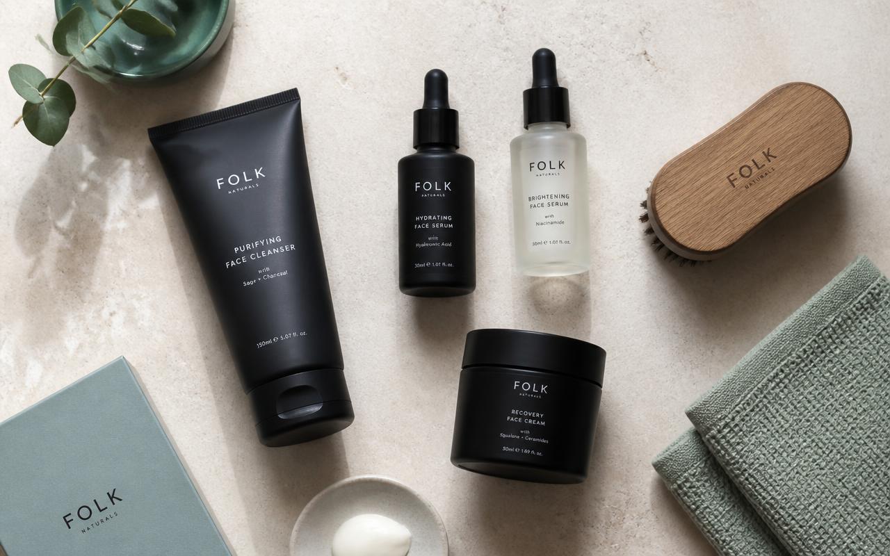 Flat-lay of men's skincare products — matte black cleanser tube, serum bottles, face cream jar, wooden grooming brush and a sage towel on a stone surface